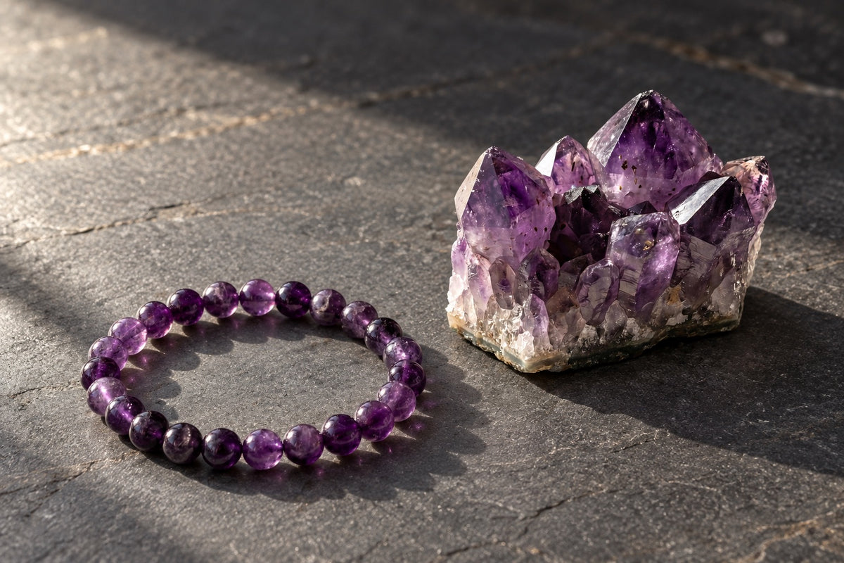 Amethyst bracelet beside a raw amethyst cluster on dark slate in warm light