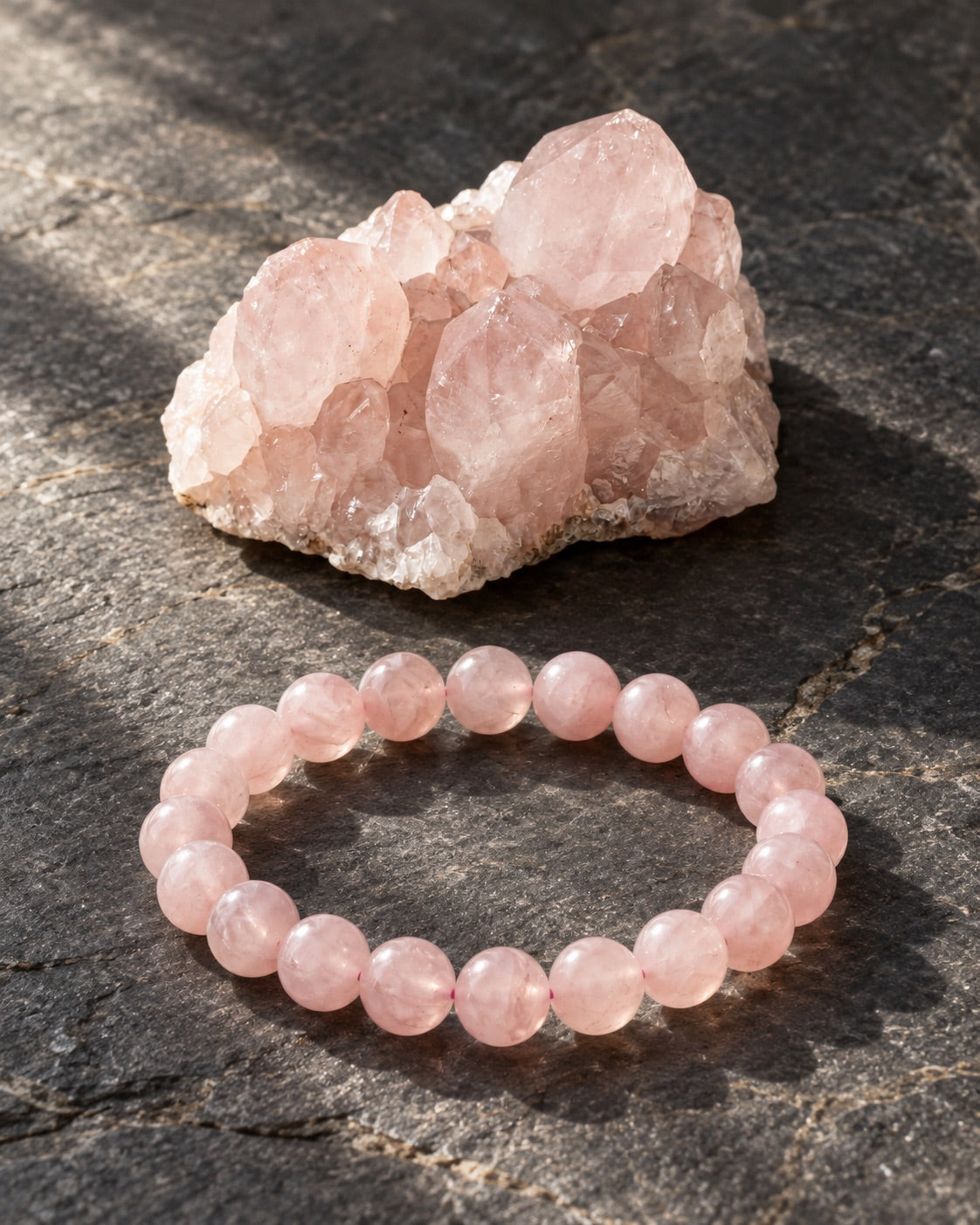 Rose quartz bracelet beside a raw rose quartz cluster on dark slate